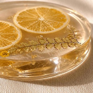 May include: A clear resin coaster with two slices of dried lemon and a fern leaf embedded inside. The coaster has a round shape and a light yellow hue, with small gold flakes scattered throughout.