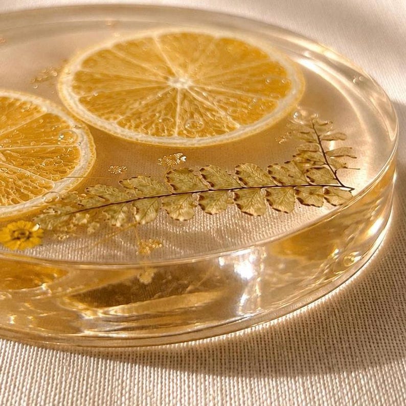 May include: A clear resin coaster with two slices of dried lemon and a fern leaf embedded inside. The coaster has a round shape and a light yellow hue, with small gold flakes scattered throughout.