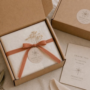 May include: A brown cardboard box with a white tissue-wrapped item tied with a rust-colored ribbon and a round sticker. The box is open, revealing the contents. A smaller box and a thank you card are also visible. The text on the card reads "Thank you for supporting handmade."