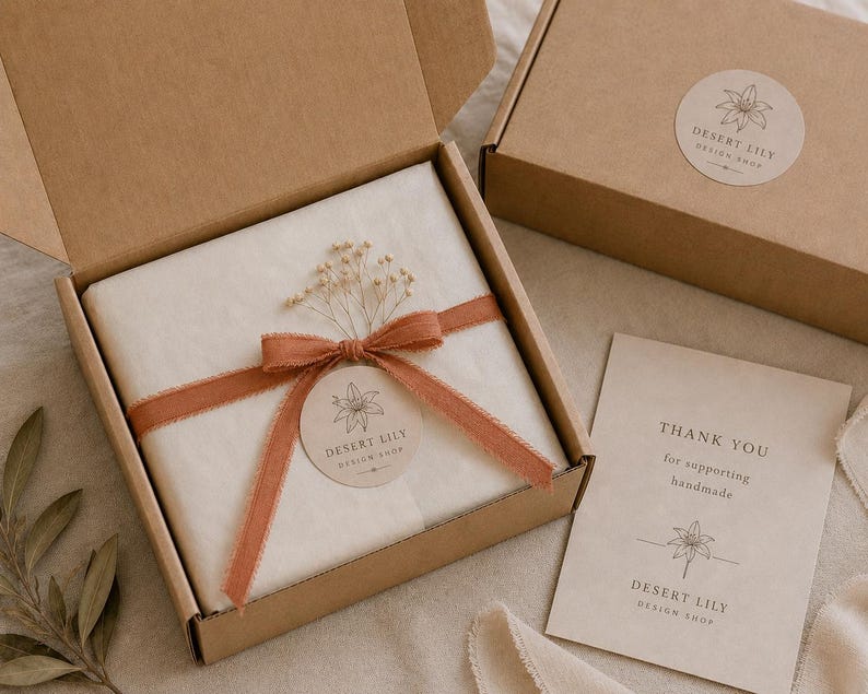 May include: A brown cardboard box with a white tissue-wrapped item tied with a rust-colored ribbon and a round sticker. The box is open, revealing the contents. A smaller box and a thank you card are also visible. The text on the card reads "Thank you for supporting handmade."