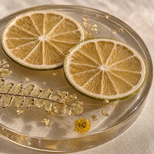 May include: A clear resin coaster with two dried lime slices, gold leaf flakes, and dried foliage. The coaster has a round shape and a glossy finish, with a light yellow and green color scheme. The design is natural and organic.