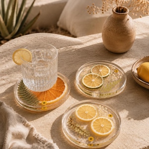May include: Clear resin coasters with embedded citrus slices and botanical elements. One coaster holds a glass of water with a lemon slice. The coasters feature orange, lemon, and lime slices, with yellow and green accents.
