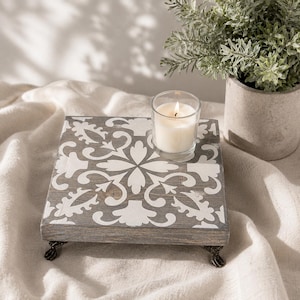 May include: A square, gray wooden riser with a white floral design. The riser has decorative metal feet and holds a lit candle in a clear glass. A potted plant sits nearby.