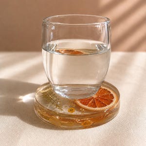May include: A clear glass of water sits atop a round resin coaster. The coaster is embedded with dried flowers, gold flakes, and a slice of dried orange. The coaster is approximately 4 inches in diameter.
