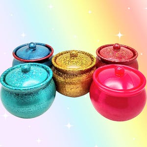 May include: Five small, round, lidded containers with a glossy finish. The collection includes a teal glitter jar, a dark blue jar with a red rim, a gold glitter jar, a red glitter jar, and a solid bright pink jar, each featuring a central knob.