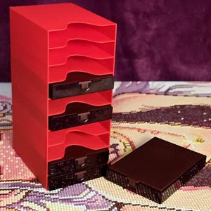 Puede incluir: Una unidad de almacenamiento vertical roja con siete ranuras abiertas, que incluye cuatro cajones rectangulares negros insertados en las secciones inferiores y un cajón negro adicional colocado por separado a la derecha de la torre.