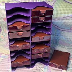 Puede incluir: Un organizador de almacenamiento de plástico morado con dos columnas verticales de ranuras. La unidad contiene varios cajones rectangulares pequeños con frentes marrones y tiradores integrados. Un cajón a juego adicional se encuentra a la derecha.