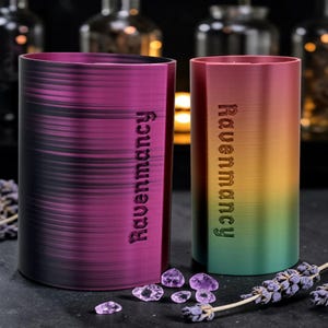 May include: Two cylindrical tumblers, one in deep purple and the other with a gradient of red, yellow, and green. The word "Ravenmancy" is vertically etched on each tumbler. Amethyst crystals and lavender sprigs are scattered around the base.