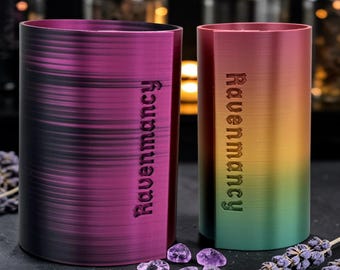 Puede incluir: Dos recipientes cilíndricos con el texto vertical Ravenmancy. El recipiente izquierdo presenta un degradado de color morado oscuro y negro, mientras que el derecho muestra un degradado de arcoíris vibrante de rojo a amarillo y verde.