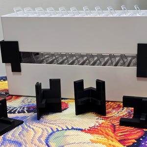 May include: A white rectangular storage container with a row of clear plastic compartments inside, featuring black corner brackets attached to the sides and four additional black L-shaped corner brackets placed in front of the container.
