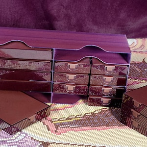 Puede incluir: Un organizador de almacenamiento morado que cuenta con una unidad principal grande con múltiples compartimentos rectangulares y varios cajones pequeños a juego. Los cajones tienen pequeñas asas claras y un acabado brillante.