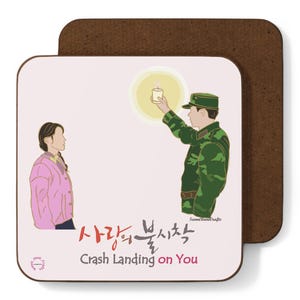 May include: Square coaster with a brown cork backing and a white front featuring a cartoon illustration. The illustration depicts two people and text in Korean and English that reads "Crash Landing on You."