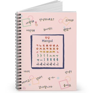 May include: A spiral-bound notebook with a light pink cover featuring Korean Hangul characters and phrases in a dark blue box. The notebook has a silver spiral binding and white lined pages. Phrases include "사랑해" and "감사합니다".