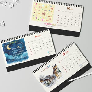 Puede incluir: Tres calendarios de escritorio con espiral y bases negras. Los calendarios muestran diseños mensuales con texto e ilustraciones coreanas. Un calendario presenta una escena de cielo nocturno, otro una casa tradicional coreana en invierno, y el tercero tiene caracteres coreanos coloridos.