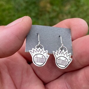 May include: A pair of silver earrings with a cartoon face design. The face has a wide mouth, two eyes, and a spiky crown.