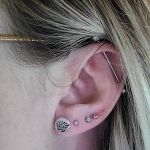 May include: A close-up of a person's ear with multiple piercings. The ear has a silver industrial barbell piercing, a silver stud with a black and white graphic, and two silver studs with clear gemstones.