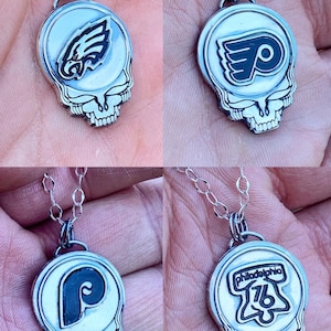 May include: Four silver necklaces with different sports team logos on the pendants. The pendants are shaped like a skull with a smiling face. The logos are the Philadelphia Eagles, the Philadelphia Flyers, the Philadelphia Phillies, and the Philadelphia 76ers.