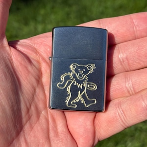 May include: A silver-colored lighter with a gold-colored dancing bear design. The lighter is rectangular and has a hinged lid. The bear is a stylized outline of a bear with its arms and legs outstretched.