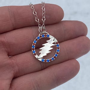 May include: Silver necklace with a circular pendant featuring a lightning bolt design surrounded by red and blue crystals.