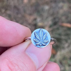 May include: A silver ring with a circular pendant displaying a detailed cannabis leaf design. The ring is held against a blurred green and brown background, highlighting the ring's intricate details and metallic sheen.