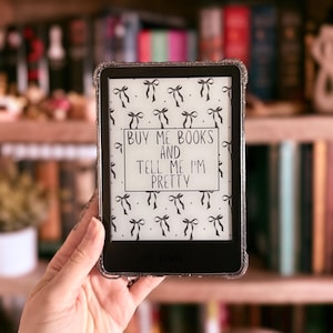 Puede incluir: Un lector electrónico Kindle en blanco y negro con un fondo blanco y lazos negros. La pantalla muestra el texto "Buy me books and tell me I'm pretty".