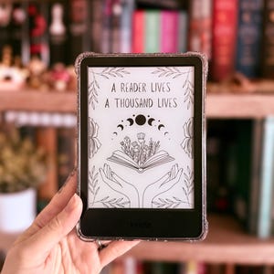 May include: A Kindle e-reader with a black and white illustration of a book with flowers and hands reaching up to it. The text on the screen reads "A reader lives a thousand lives".