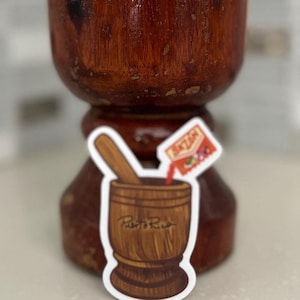 May include: A wooden mortar and pestle with a sticker of a mortar and pestle with the text "Puerto Rico" and a red and white packet of seasoning with the text "Sazon" on it.
