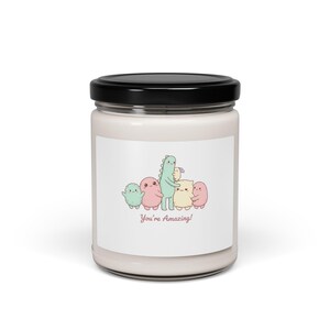 Peut inclure: Un pot à bougie blanc avec un couvercle noir. L'étiquette présente une illustration de dessin animé de plusieurs créatures colorées, dont un dinosaure, se tenant dans les bras. Le texte "You're Amazing!" est imprimé en dessous. Une bougie décorative.
