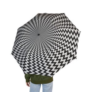 Puede incluir: Paraguas abierto con un patrón de zigzag en blanco y negro. El diseño presenta un patrón de chevrón repetido, creando un efecto de vórtice visual. La persona que lo sostiene lleva una chaqueta verde y vaqueros azules.