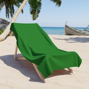 May include: A bright green beach towel on a wooden deckchair on a sandy beach. The towel is a solid colour. In the background, there's a boat and the sea under a clear blue sky.