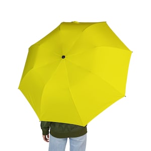 May include: Bright yellow umbrella, open and displayed against a white background. The umbrella's canopy is a vibrant, solid yellow color. The umbrella is held by a person wearing a green jacket and blue jeans.