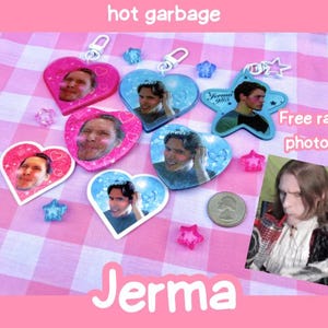 May include: A collection of heart and star-shaped acrylic keychains featuring photographic portraits of a person, accompanied by small star-shaped beads and a US quarter coin. A rectangular card shows a person drinking from a red cup.