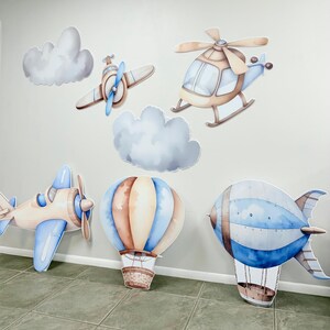 May include: A set of six blue and brown watercolor-style cutouts of various modes of transportation, including a hot air balloon, a blimp, a helicopter, and two airplanes. The cutouts are perfect for a baby shower or birthday party.
