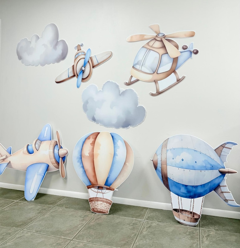 May include: A set of six blue and brown watercolor-style cutouts of various modes of transportation, including a hot air balloon, a blimp, a helicopter, and two airplanes. The cutouts are perfect for a baby shower or birthday party.