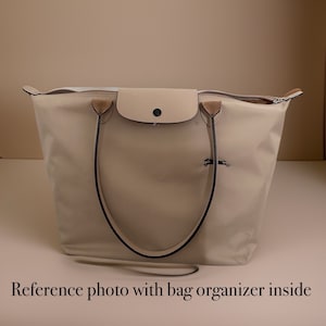 May include: A beige tote bag with black handles and a small beige flap pocket on the front. The bag has a zipper closure and a small horse logo on the front.