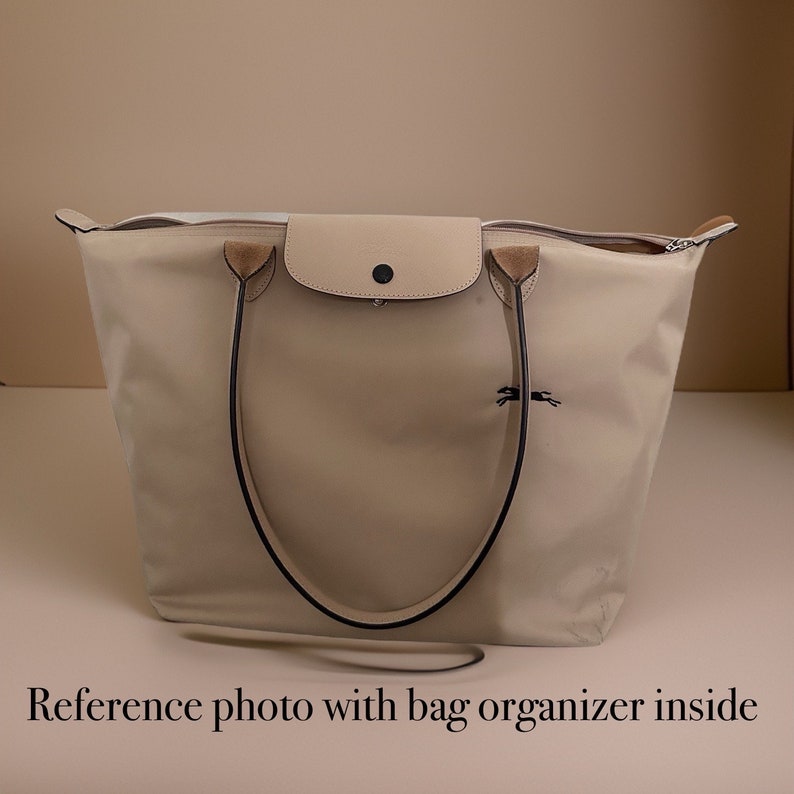 May include: A beige tote bag with black handles and a small beige flap pocket on the front. The bag has a zipper closure and a small horse logo on the front.