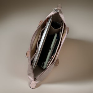 May include: A light pink tote bag with multiple interior pockets. The bag is open and shows a laptop, a phone, and a notebook inside.