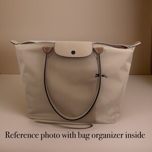 May include: A beige tote bag with black handles and a small beige flap pocket on the front. The bag has a zipper closure and a small horse logo on the front.