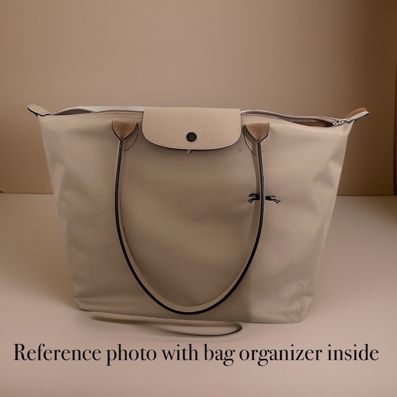 May include: A beige tote bag with black handles and a small beige flap pocket on the front. The bag has a zipper closure and a small horse logo on the front.