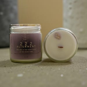 May include: Two glass jar candles with white wax. One jar has a label with the text "222 ALIGNMENT" and "YOU ARE AT THE RIGHT PLACE AT THE RIGHT TIME". The other candle has a wooden wick and a small crystal on top. The candles are 2.5 oz.