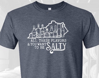 May include: A dark heather grey t-shirt with a white outline of the state of Kentucky and a row of liquor bottles. The text "All these flavors & you want to be Salty" is printed on the shirt.