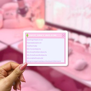 May include: A rectangular sticker with a light pink border and white background featuring the text CHEAT CODES-BUILDING at the top, followed by a list of game commands including testingcheats true, motherlode, and bb.moveobjects in purple font.