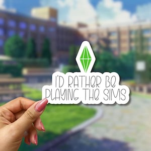 May include: A white die-cut sticker featuring a green diamond-shaped plumbob icon above the black text that reads I'D RATHER BE PLAYING THE SIMS in a rounded, whimsical font. The sticker has a thin white border and a matte finish.