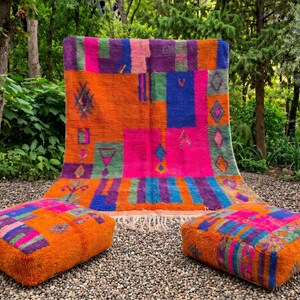 May include: A large rectangular area rug featuring a vibrant patchwork of orange, hot pink, purple, blue, and green blocks with geometric diamond and linear patterns. Two matching square floor cushions display the same colorful, textured design.
