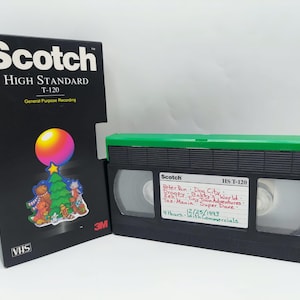 Può includere: Una videocassetta VHS nera Scotch High Standard T-120 e la sua scatola. La scatola presenta il logo Scotch e una grafica colorata di un albero di Natale e orsacchiotti. Il nastro ha una parte superiore verde e un'etichetta con testo manoscritto.