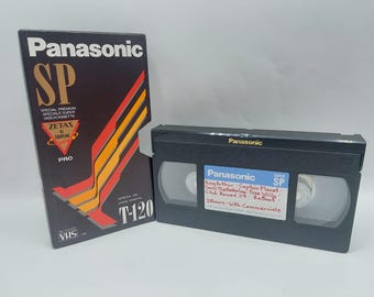 Pode incluir: Uma cassete de vídeo VHS Panasonic T-120 preta com a sua capa de cartão correspondente. A capa apresenta riscas diagonais vermelhas e cor de laranja e o texto Panasonic SP. A cassete tem uma etiqueta branca com texto manuscrito listando títulos.