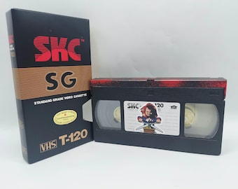Könnte beinhalten: Eine schwarze SKC SG T-120 Standard-Videokassette mit passender Pappschachtel. Die Schachtel zeigt roten Text und ein Good Housekeeping-Siegel. Die Kassette hat ein weißes Etikett mit einer Grafik einer Puppe, die eine Schere hält.