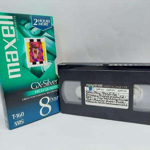 Könnte beinhalten: Eine Maxell VHS-Videokassette und ihre Box. Die Box ist türkis und weiß mit den Wörtern "GX-Silver" und "8 Stunden" bedruckt. Die schwarze Videokassette hat ein weißes Etikett mit handschriftlichem Text.