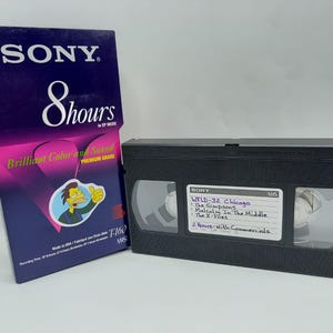 Può includere: Una videocassetta VHS Sony nera e la sua custodia. La custodia è blu scuro con la scritta "SONY 8 hours" e un'immagine a cartoni animati. L'etichetta del nastro elenca programmi TV come "The Simpsons".