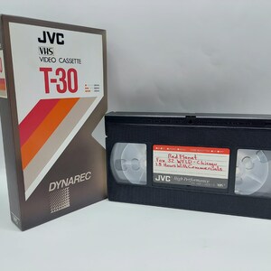 Op de afbeelding: Een JVC T-30 VHS-videocassette in de originele bruin-witte doos met oranje en rode diagonale strepen. Daarnaast ligt een zwarte VHS-band met een wit label waarop handgeschreven tekst staat: Red Planet, Fox 32 WFLD-Chicago, 1.5 Hours.